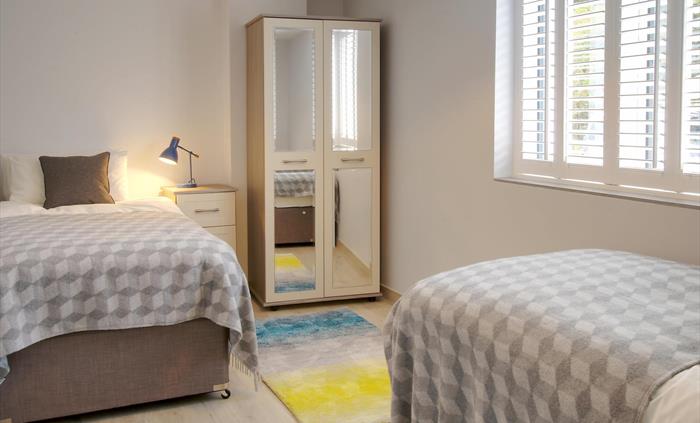 Twin bedroom with geometric throws, fresh white walls, and pops of colour from green lamps and fresh daffodils.